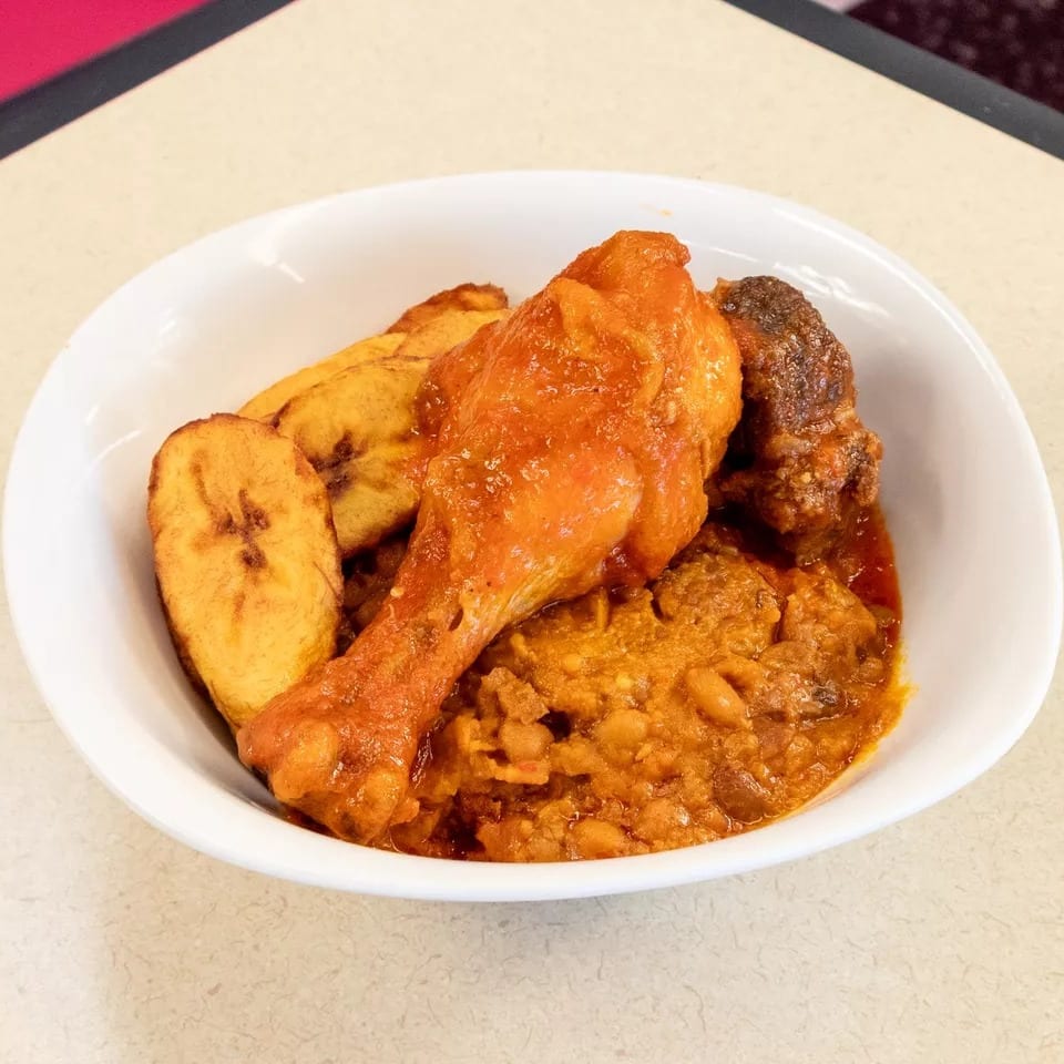 A white bowl filled with chicken, plantains, and a rich stew on a light-colored table.